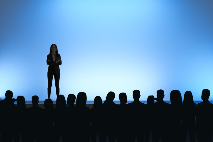 BusinesswBusinesswoman giving speech in front of backlit audience on light blue background. Speaker and leader conceptoman giving speech in front of backlit audience on light blue background. Speaker and leader concept